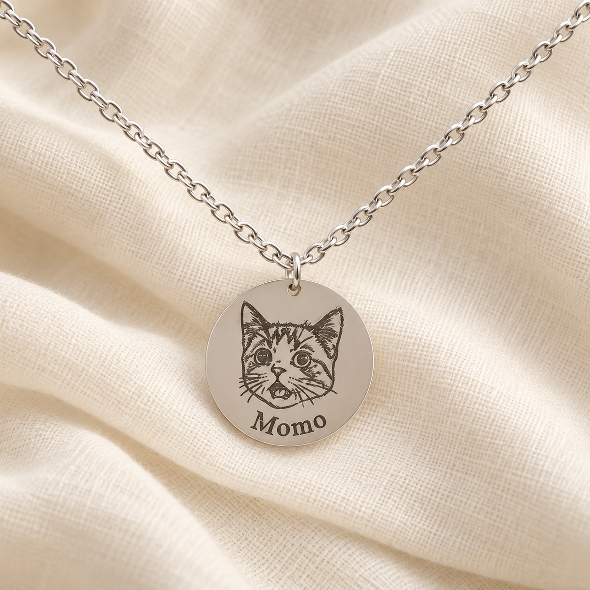 Silver-tone Momo cat portrait necklace on soft ivory linen; styled image